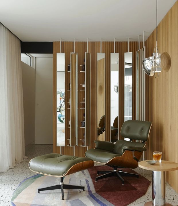 A refined lounge or reading area with two olive-green leather lounge chairs and matching ottomans, set against a vertical timber-panelled feature wall. Slim suspended mirrored shelving and display panels create rhythm along the wall, while a sculptural chrome-and-glass pendant light hangs beside a small round side table. The room has a pale terrazzo floor, sheer curtains, neutral walls, and layered geometric rugs. The overall look is sophisticated mid-century-inspired with contemporary detailing.
