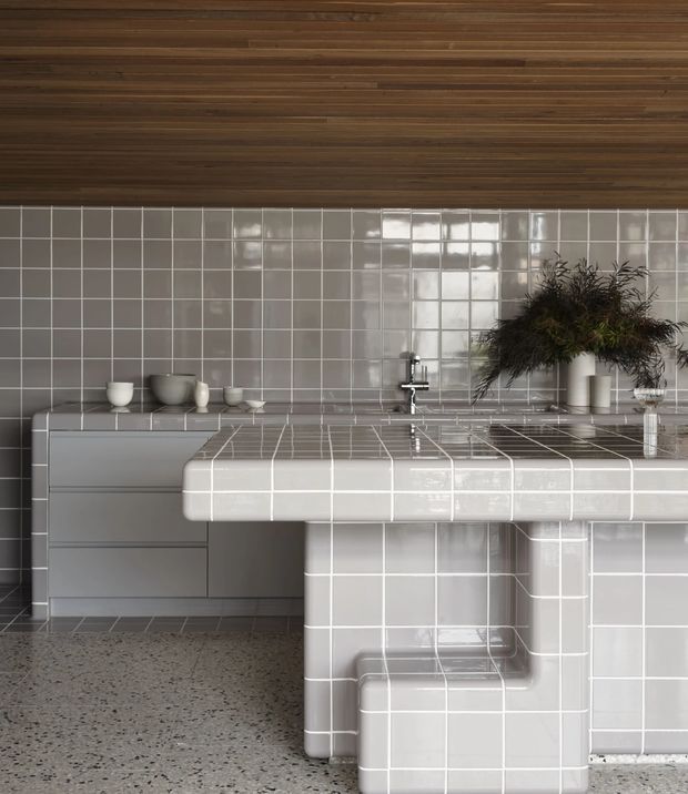 A refined contemporary kitchen featuring a fully tiled island and backsplash in glossy light-grey square tiles, integrated cabinetry, a stainless-steel tap, terrazzo-style flooring, and a warm timber-lined ceiling. Minimal ceramic vessels and foliage add understated styling.