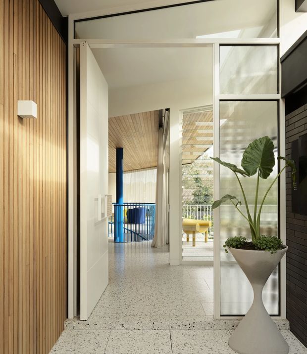 A bright modern entryway or hallway with a terrazzo floor, warm vertical timber wall panelling, white walls and framed translucent glass partitions. A sculptural white planter with a large tropical plant stands beside a dark brick feature wall. Through the glazed partition, a blue-railed stair or landing area and additional timber-lined surfaces are visible.
