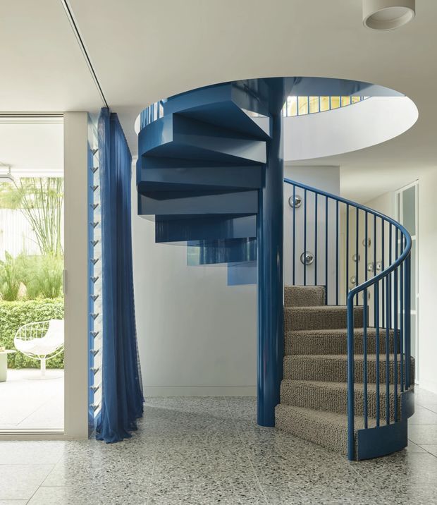A bright contemporary interior focused on a sculptural blue spiral staircase with matching curved balustrade and central support. The staircase rises through a large oval opening in the ceiling, with a glimpse of an upper-level landing. Pale walls and ceiling, terrazzo flooring, and carpeted stair treads create a clean, graphic setting. Blue curtains and a glazed opening connect the stair hall to a planted outdoor area.
