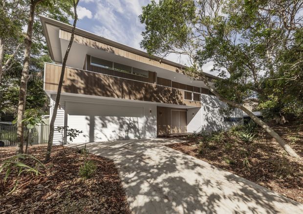 Exterior photograph of a contemporary multi-level house set among mature trees. The home has a prominent sloping roof, white rendered walls, warm timber-toned balcony screening and entry cladding, a large enclosed garage, and a concrete driveway leading toward the street. Dense landscaped vegetation surrounds the facade.