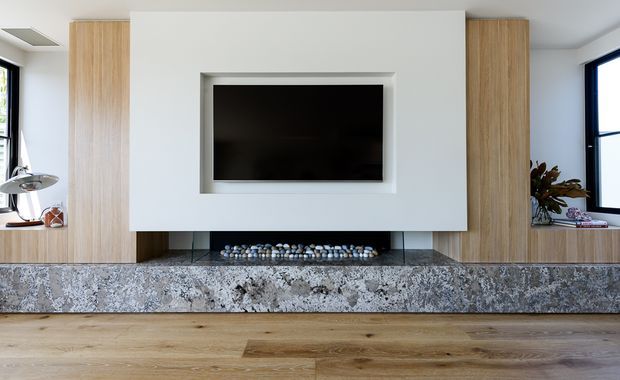 A contemporary living room media wall with a centrally mounted television, pale timber vertical cabinetry on both sides, and a long textured stone fireplace hearth with a recessed firebox filled with decorative stones. Warm timber flooring, neutral walls, black-framed windows, and minimal furnishings create a clean, refined composition.