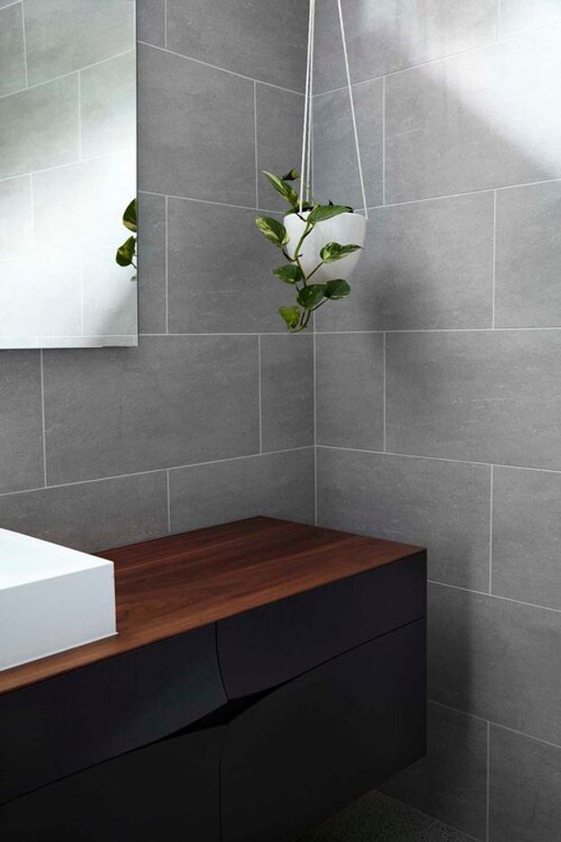 A compact contemporary bathroom corner with large grey wall tiles, a dark matte vanity with sculptural drawer fronts, and a warm dark timber benchtop. A white vessel basin is partially visible beside a wall mirror, while a hanging potted vine adds greenery.