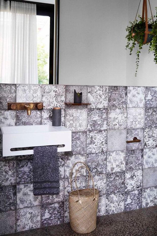 A compact, stylish bathroom vanity area featuring a wall-mounted white basin with a dark towel, copper-toned wall-mounted tapware, and a full-height patchwork backsplash of ornate grey, white, and charcoal patterned tiles. A black-framed window with sheer curtains brings in natural light, while a hanging trailing plant and woven basket add texture. The floor appears to be dark terrazzo or pebble-textured concrete.