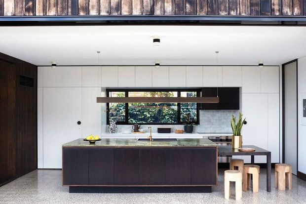 A contemporary open-plan kitchen viewed straight on, with a large dark timber island, pale stone benchtop, white full-height cabinetry, integrated appliances, a horizontal window, and a white brick-pattern splashback. A dark linear pendant hangs above the island, while a black dining table and stools sit to the right. The floor is a polished terrazzo-like finish, and dark timber cladding is visible along the upper edge and side wall.