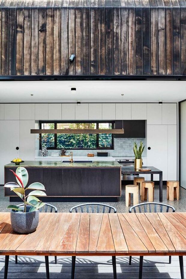 A contemporary open-plan kitchen and dining area with a dark timber island, white full-height cabinetry, pale tiled splashback, and a large horizontal window overlooking greenery. A long timber dining table with black-framed chairs occupies the foreground, while a raised timber-clad ceiling adds warmth and texture. Bar stools, integrated appliances, pendant-style task lighting, and indoor plants complete the space.