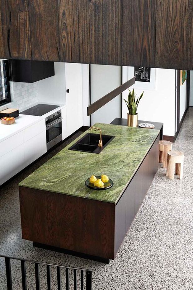 Contemporary kitchen viewed from an elevated angle, centred on a large island with a dramatic green veined stone benchtop, integrated black sink and brass tap. The island and overhead feature cabinetry are clad in dark timber, contrasting with white cabinetry, white tiled splashback and black appliances. Terrazzo flooring, timber stools, a potted plant and concealed extraction detail complete the space.