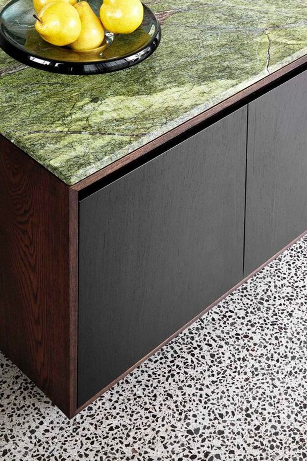 Close-up product-style image of a dark timber and charcoal cabinet or island with a distinctive green veined stone benchtop. The unit sits on a black-and-white terrazzo floor, with a black glass fruit bowl containing pears on the benchtop. The composition highlights the material contrast and crisp joinery details.