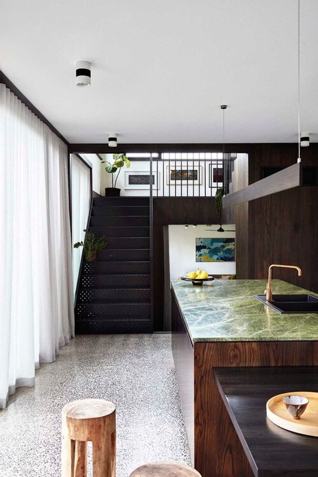 A sophisticated open-plan kitchen and stair area with dark timber wall panelling and joinery, a green-veined stone island benchtop, black undermount sink, and warm brass tapware. A perforated dark stair rises beside full-height sheer curtains, with a glazed internal balustrade and artwork beyond. Terrazzo-style flooring, pendant and surface-mounted lighting, indoor plants, and timber stools complete the refined interior.