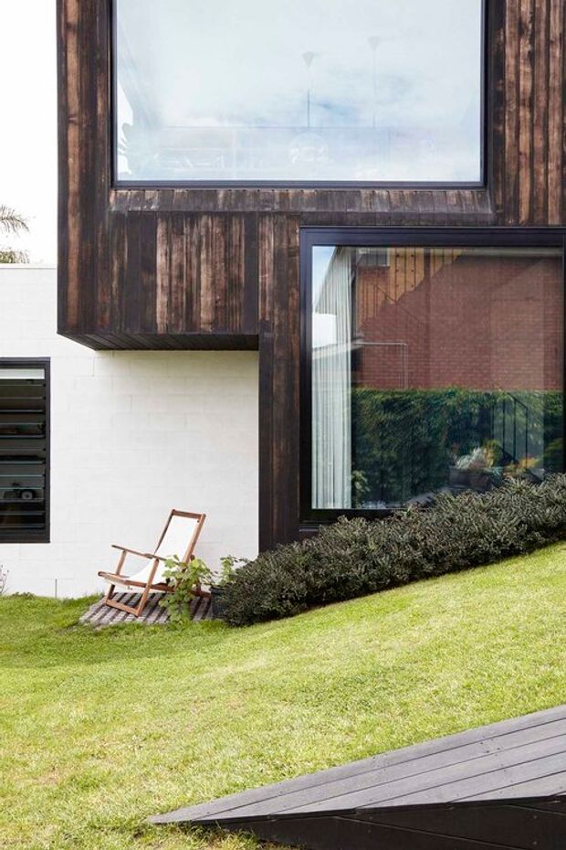 Exterior view of a contemporary home with dark vertical timber cladding projecting around large black-framed windows, contrasting white masonry walls, and a landscaped sloping lawn. A timber deck or ramp occupies the foreground, with low shrubs and an outdoor timber lounge chair beside the facade.
