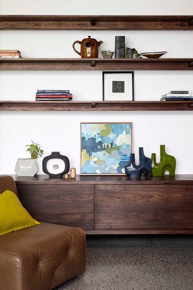 A styled living room vignette featuring dark timber floating shelves and a matching low sideboard against a white wall. The shelves display books, ceramics, a vintage-style teapot, framed artwork and decorative objects. A brown leather sofa with a bright yellow cushion occupies the foreground, with a speckled grey floor and colourful artwork and vases adding visual interest.