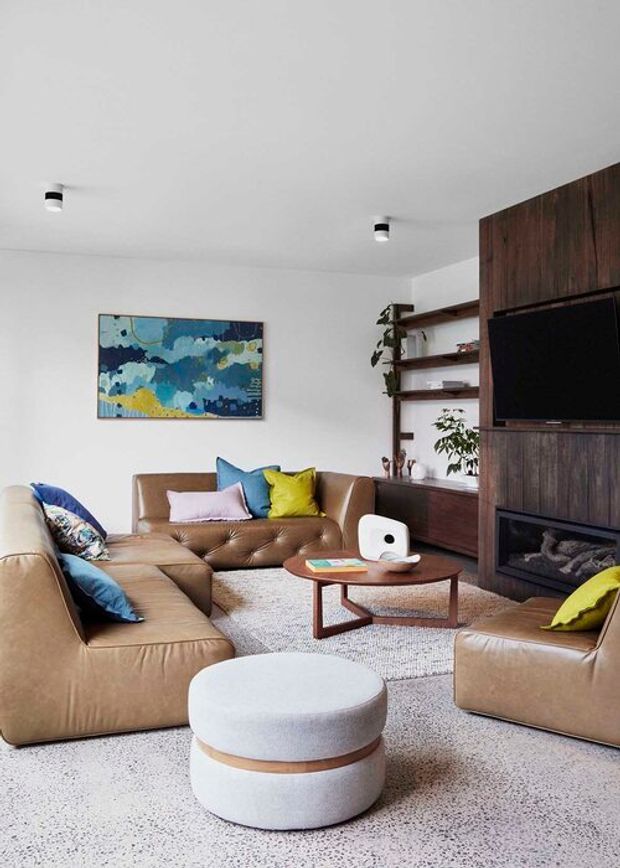 A bright, contemporary living room with generous tan leather sofas arranged around a low wooden coffee table. The room features a large textured terrazzo-like floor, a pale upholstered round ottoman, colourful cushions, abstract artwork, open timber shelving, and a dark timber built-in media wall with a television and fireplace.