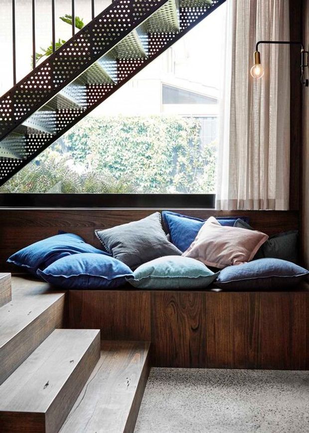 Interior view of a contemporary living area with a dark timber built-in window seat layered with blue, grey, and blush cushions. Pale timber stairs rise beside the seating, while a perforated dark metal stair balustrade crosses the upper portion of the image. Large glazing brings in abundant daylight, with full-height neutral curtains and greenery visible outside. A slim black wall light adds a warm accent.