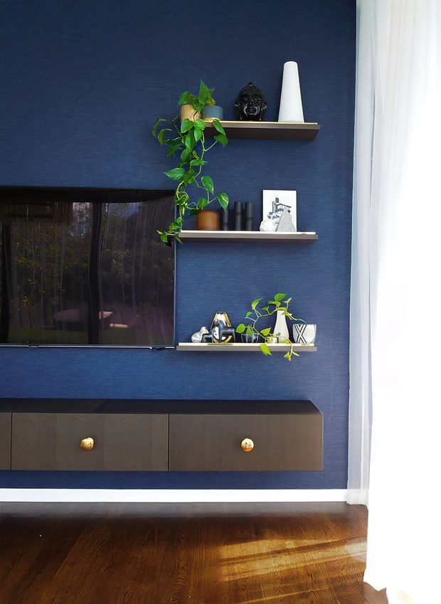 A styled living room media wall with deep navy textured wall finish, a wall-mounted television, dark timber floating media cabinetry with brass knobs, and three floating display shelves. The shelves hold plants, pottery, sculptural objects, books, and artwork. White curtains at the right allow bright natural light into the room, contrasting with the dark stained timber floor. The overall look is refined, contemporary, and moody.