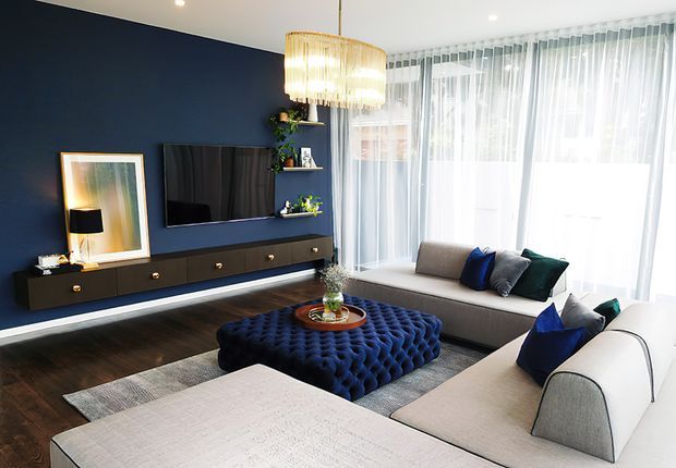 A bright, carefully styled contemporary living room with a deep navy feature wall, wall-mounted television, floating dark timber media cabinetry, and a large cream sectional sofa arranged around a deep blue tufted ottoman. Floor-to-ceiling sheer curtains provide abundant natural light, while a large woven pendant adds warmth above the seating area. Dark timber flooring, a neutral rug, and blue accent cushions complete the refined, high-contrast scheme.