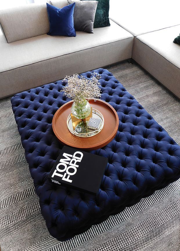 Close-up interior view of a refined living room seating arrangement. A light neutral L-shaped upholstered sofa surrounds a large deep navy button-tufted ottoman, topped with a round timber tray table, glass vase and decorative accessories. A monochrome patterned rug covers the floor, while navy, grey and teal cushions add contrast. The styling has a polished, contemporary feel with luxurious textile details.