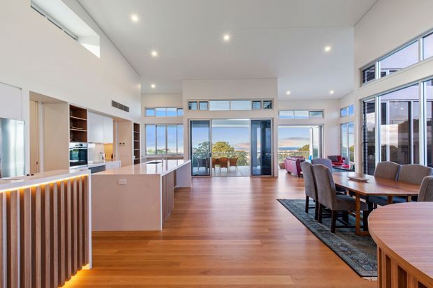 Bright, open-plan contemporary living space combining a kitchen, dining area and lounge. The room features warm timber flooring, pale built-in cabinetry, a large central island, extensive floor-to-ceiling glazing, and a high white ceiling. Water views are visible beyond the glazing, while a timber dining table and upholstered chairs occupy the foreground.