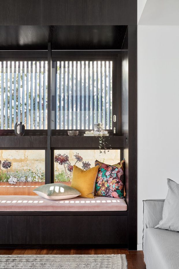 A bright, refined interior featuring a built-in dark timber window seat with a pink upholstered cushion, colourful patterned and mustard pillows, and vertical blinds filtering sunlight. The seat incorporates deep timber cabinetry and display shelving, with a white upholstered sofa partially visible at right, timber flooring, and a pale patterned rug.