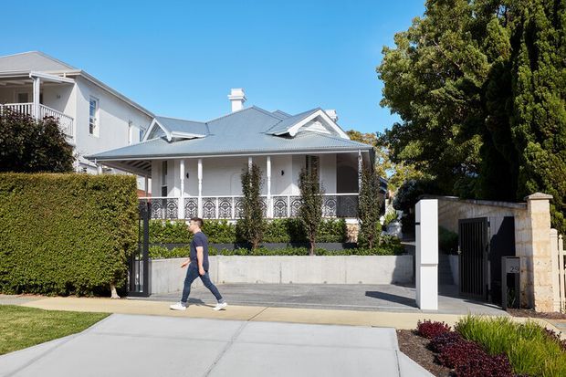 Street-facing view of a light-coloured, single-storey residential house with a broad hipped metal roof, covered front veranda, decorative metal balustrades, white columns, and landscaped garden beds. A driveway and gated side carport or garage entry are visible, with a larger adjoining or neighbouring dwelling behind. The scene is bright and suburban with established hedges and mature trees.