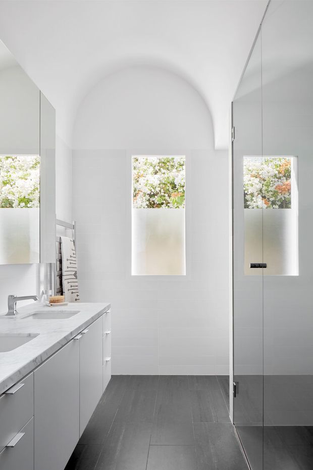 Bright contemporary bathroom with a white tiled wall, arched ceiling detail, two narrow frosted-glass windows, a large mirrored wall cabinet, and a pale stone vanity with integrated basins. A frameless glass shower enclosure occupies the right side, while dark grey large-format floor tiles provide contrast. Garden foliage is visible beyond the windows.