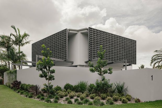 Contemporary tropical residential exterior with a screened upper volume wrapped in patterned breeze-block masonry, supported over a recessed lower level. The building has a restrained grey and white palette, flat rooflines, deep overhangs, and a landscaped front garden with palms and drought-tolerant planting.