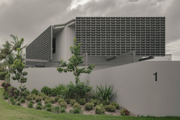 Exterior view of a contemporary multi-level residence with a large patterned breeze-block screen façade, corrugated metal wall sections, projecting roof or awning elements, and a restrained grey palette. The home sits behind a rendered boundary wall marked with the number 1 and is fronted by landscaped planting and lawn under an overcast sky.
