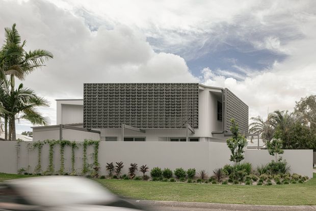 Exterior view of a contemporary multi-level residence behind a low rendered boundary wall. The house features strong geometric volumes, broad perforated breeze-block screens, flat roof forms, and pale neutral finishes. Tropical palms, landscaped planting, and a suburban street frontage frame the building.