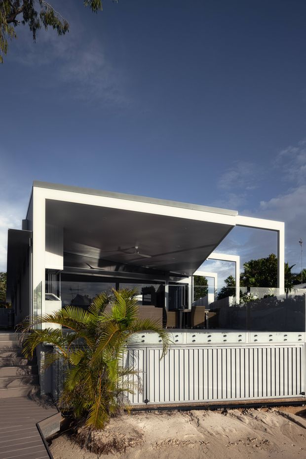 Modern two-storey-looking beachfront residence with a bold white rectangular architectural frame, deep covered outdoor living area, dark glazed walls, and a projecting flat roof. The house sits immediately beside sandy ground, with palms, a raised timber deck, glass balustrades, and a white metal boundary fence visible in the foreground. The image is an exterior architectural photograph under a clear blue sky.