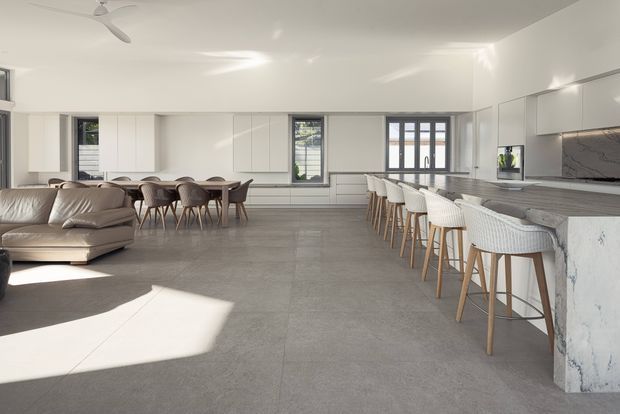 A spacious open-plan contemporary interior combining a kitchen, dining area and living room. The room features a long island with white textured bar stools, extensive white cabinetry, a grey stone-look benchtop and splashback, large-format grey flooring, a dining table with upholstered chairs, a pale sectional sofa, expansive glazing and a ceiling fan. Bright natural light creates a clean, airy atmosphere.