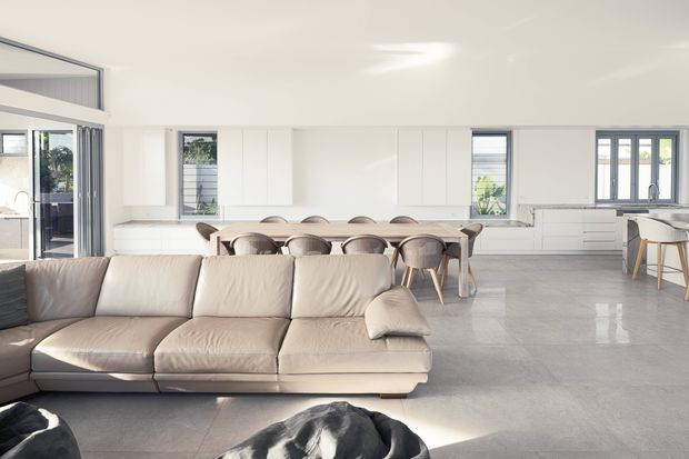 Bright open-plan interior combining a living room, dining area and kitchen. A large cream sectional sofa occupies the foreground, with a timber dining table and upholstered chairs behind it. The kitchen has white full-height cabinetry, pale benchtops and a large island or peninsula, while expansive glazing and a polished grey floor create an airy, contemporary feel.