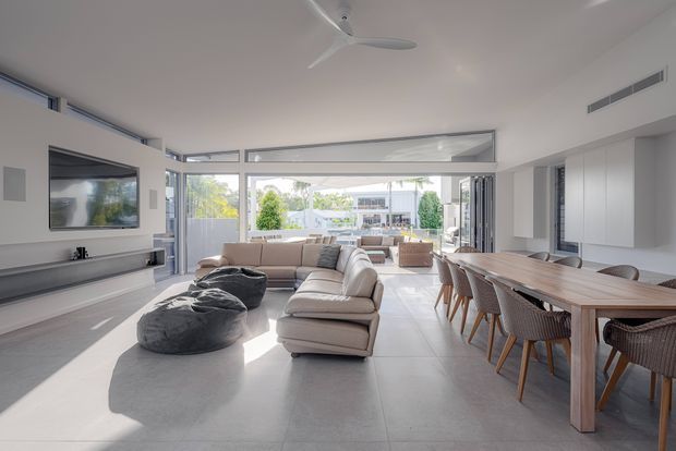 Bright open-plan contemporary living and dining area with floor-to-ceiling glazing opening toward an outdoor terrace. The room features a large sectional sofa, wall-mounted television and streamlined media joinery on the left, a substantial timber dining table with woven chairs on the right, pale tiled flooring, white walls and ceiling, and abundant natural light. A kitchen or built-in cabinetry zone is visible along the right wall.