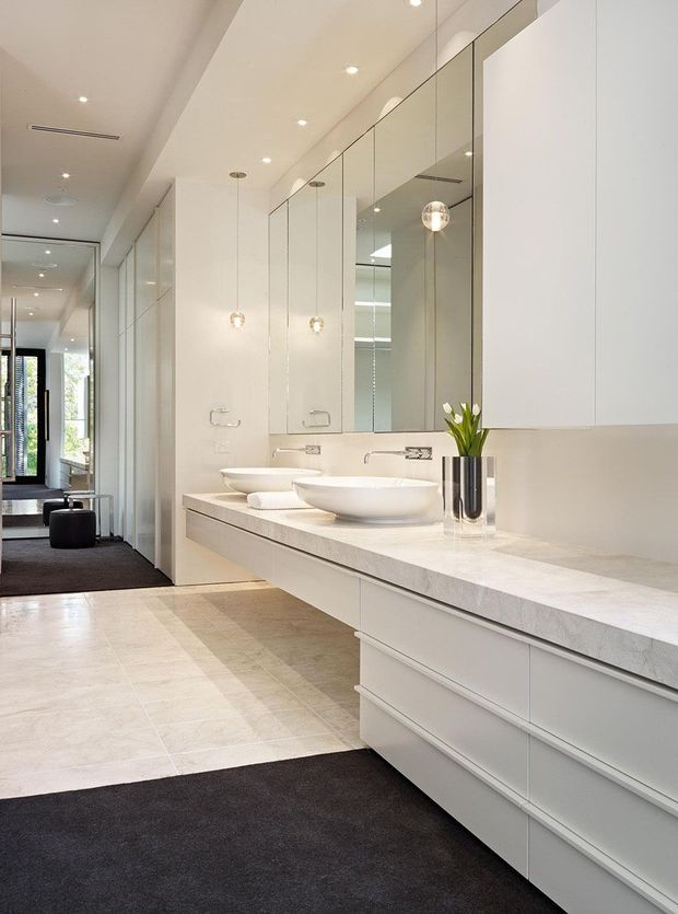 A bright, elongated contemporary bathroom with a long white vanity, multiple integrated drawers, two vessel basins, a pale stone benchtop, full-height mirrored cabinetry, and minimalist pendant lighting. Large pale floor tiles contrast with dark charcoal carpeted areas, while a corridor and glazed doorway extend into the background.
