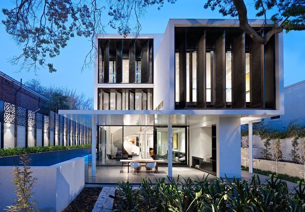 Contemporary two-storey residence with a crisp white rendered facade, extensive floor-to-ceiling glazing, projecting upper-level volumes, deep vertical sun-shading screens, and a transparent ground-floor living area. The house is set behind landscaped planting and a low wall, with a narrow blue-toned water feature or pool along the side. The composition is photographed at dusk beneath mature trees.