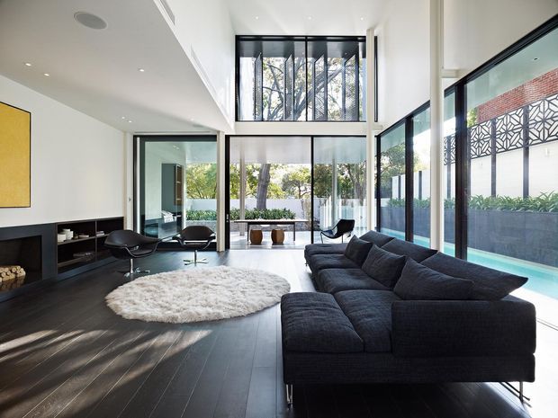 A bright, double-height contemporary living room with floor-to-ceiling glazing, a large dark sectional sofa, dark timber flooring, a minimalist fireplace and built-in shelving. The space opens toward a landscaped courtyard and features a sculptural patterned screen visible outside.