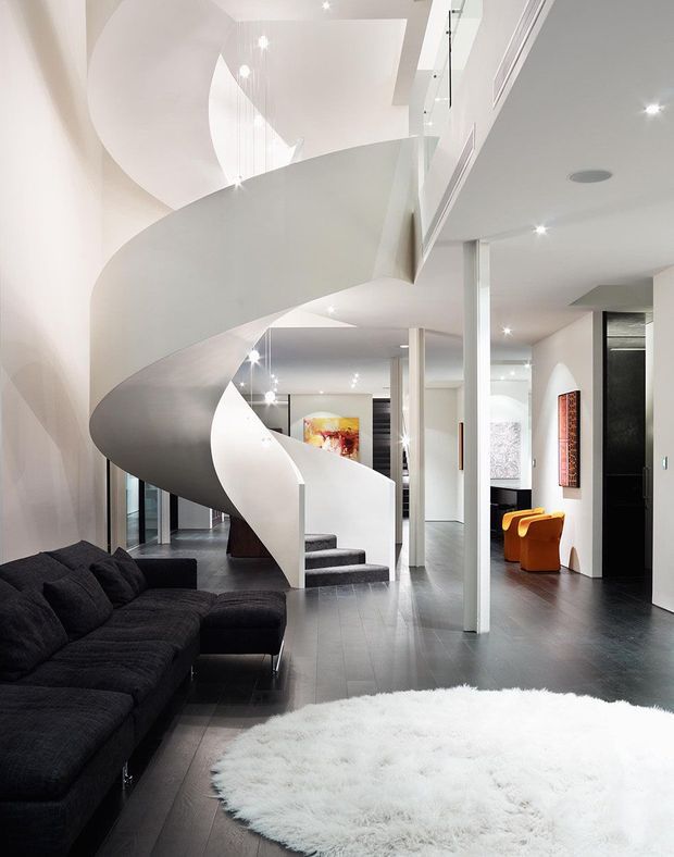 Bright, high-end contemporary interior centred on a sculptural white curved staircase with glass balustrading. The open-plan space includes a dark sectional sofa, dark timber or stained flooring, white walls and ceilings, recessed lighting, colourful accent chairs, and modern artwork.