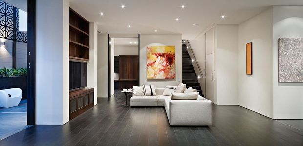 Contemporary open-plan living room with a large light sectional sofa, dark timber flooring, built-in dark timber media joinery, white walls, recessed ceiling lighting, and a staircase leading to an upper level. A glimpse of an adjacent blue-toned bathroom or feature space is visible at left, with artwork and a mirrored panel adding visual interest.