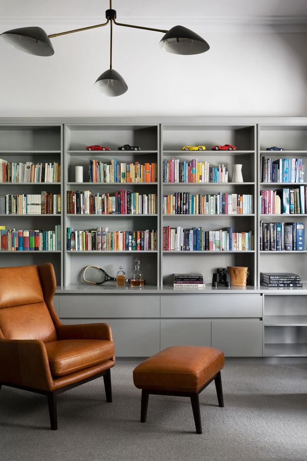 A refined living room or reading space featuring a full wall of grey built-in shelving and cabinetry filled with books and small decorative objects. A cognac-brown leather armchair and matching ottoman sit on a grey carpet, beneath a sculptural three-arm pendant light with dark metal arms and smoked glass shades. The room has a calm, sophisticated, book-lined atmosphere with clean contemporary detailing.