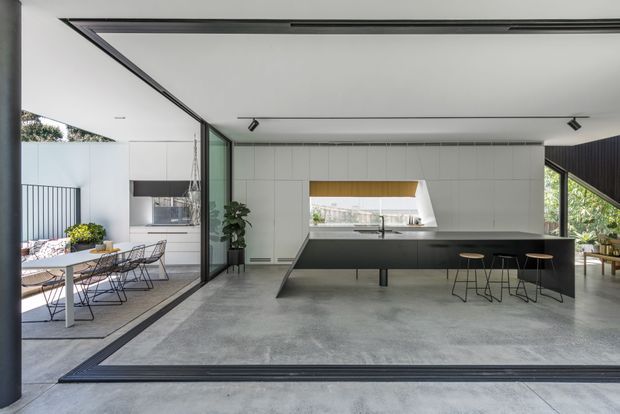 Open-plan contemporary kitchen and dining space with a minimalist, monochromatic fit-out. The kitchen features a long matte-black island with integrated sink and seating, white full-height tiled or panelled walls, a sculptural horizontal window, dark-framed glazing and doors, and a polished concrete floor. A dining area and planted courtyard are visible through the large glazed opening.