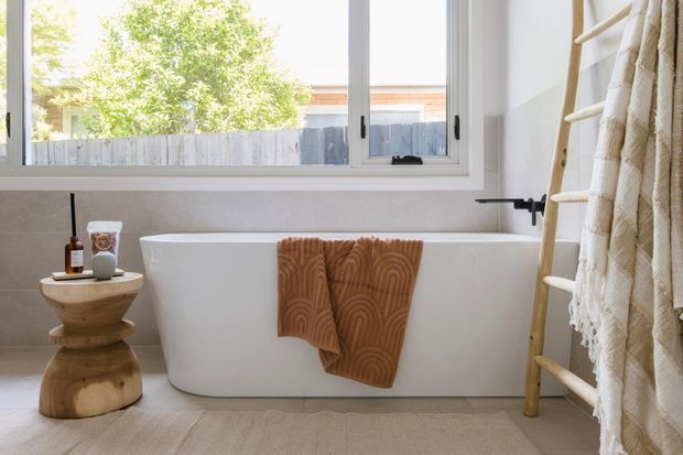 Bright, calm bathroom featuring a freestanding white bathtub beneath a wide white-framed window. Neutral textured wall and floor finishes are paired with a black wall-mounted tap, a sculptural timber side stool, and a timber ladder-style towel rack with cream textiles. The view outside is secondary.