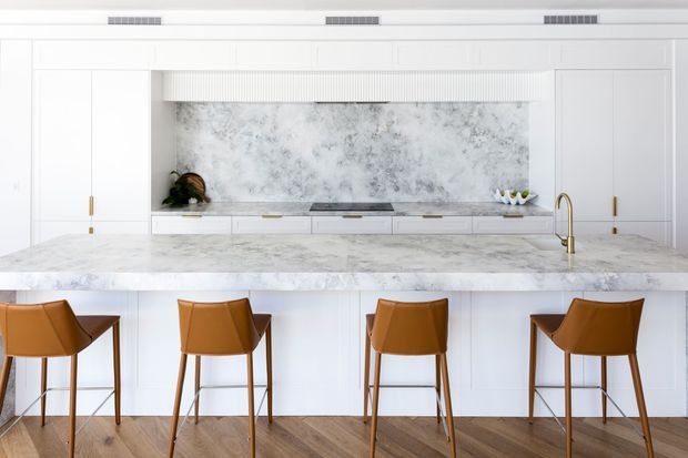 Bright, refined contemporary kitchen featuring a large marble-look island with four caramel leather bar stools, white shaker-style cabinetry, a marble-look splashback, integrated appliances, and brushed brass tapware and hardware. Warm timber flooring adds contrast to the predominantly white palette.