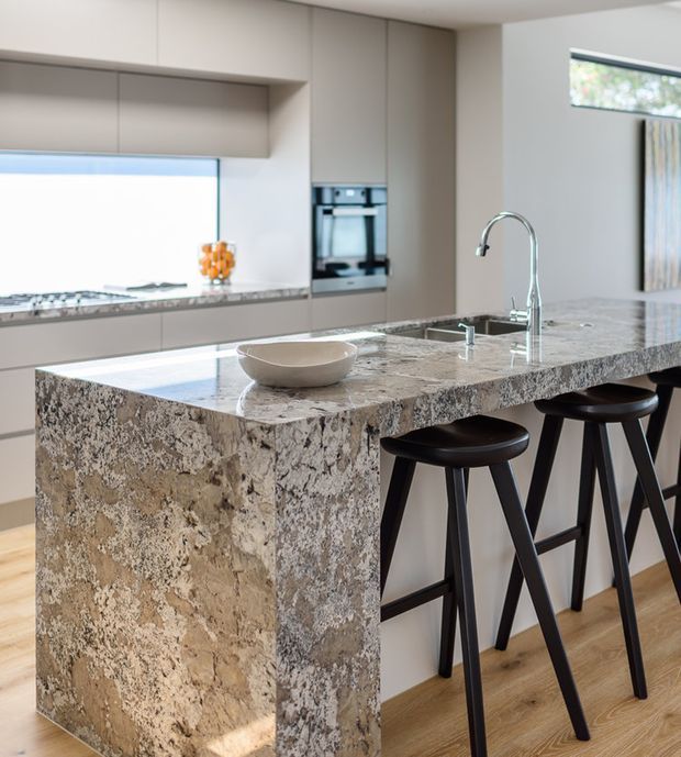 Contemporary kitchen focused on a large waterfall-edge stone island with an integrated sink and tall curved faucet. The island provides seating for three on black bar stools, while surrounding cabinetry is finished in a warm neutral tone with built-in appliances. Light timber flooring and a bright, refined palette complete the space.