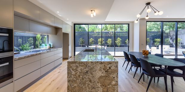 Contemporary open-plan kitchen and dining area with extensive light cabinetry, integrated appliances, a large natural-stone island, timber flooring, and black-framed floor-to-ceiling glazing opening to a landscaped courtyard. A dark dining table with upholstered chairs and sculptural pendant lighting occupy the adjacent dining zone.
