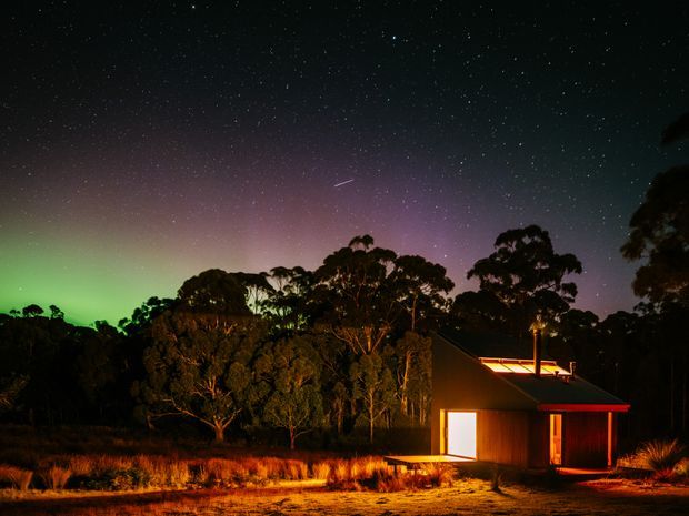 A small contemporary cabin-like house set in a wooded natural landscape at night. Warm interior and exterior lighting illuminate the dark timber-toned facade and surrounding grasses, while a star-filled sky with vivid green and purple aurora-like colours forms the backdrop. The simple building has a steep sloping roof and a compact, single-storey form.