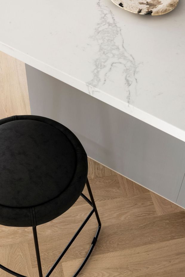 Close-up interior detail of a refined kitchen or kitchenette, showing a white marble-look stone benchtop with grey veining, light timber cabinetry and flooring, and a black upholstered bar stool with slender black metal legs. The composition focuses on material and finish details with a warm, minimalist palette.