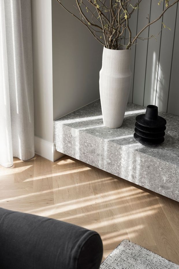 A softly lit contemporary interior vignette featuring a floating grey stone console or hearth with a large white textured vase containing branches and a black sculptural vessel. Sheer curtains, grey wall panelling, pale timber herringbone flooring, and partial black upholstered seating create a refined, minimalist atmosphere.