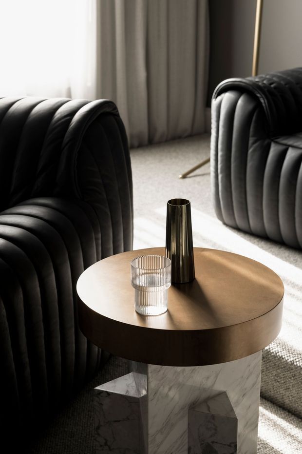 A moody, refined living room vignette featuring two black vertically channelled upholstered lounge chairs or sofas, a round bronze-toned side table with a marble pedestal base, and minimalist metallic and ribbed glass accessories. Soft daylight filters through curtains onto a neutral carpet, creating strong light-and-shadow contrast.