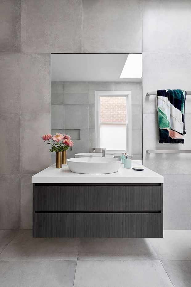 A contemporary bathroom vanity with a white engineered-stone countertop, two round vessel basins, dark vertically textured cabinetry, and a wall-mounted mirror. Large-format grey tiles cover the walls and floor, with a narrow window, recessed wall niche, chrome tapware, and a towel rail with a patterned towel. The space is clean, minimalist, and softly lit.