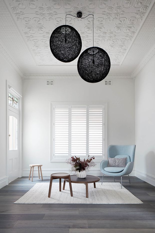 A bright, refined sitting room or entry lounge with white walls, ornate pressed-metal patterned ceiling, decorative cornices, plantation shutters, and a glazed panel door. Two large black woven pendant lights hang overhead. A pale blue upholstered armchair, round dark timber coffee tables, a neutral textured rug, and a small stool furnish the room. Dark wide-plank timber flooring contrasts with the predominantly white interior.