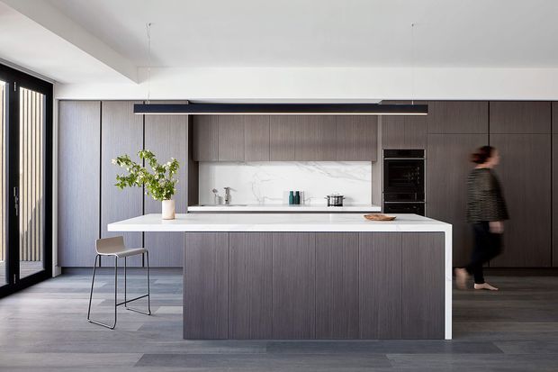 Contemporary kitchen with a large central island, dark vertically grained timber cabinetry, white stone benchtops, and a marble-look splashback. Full-height dark cabinetry incorporates integrated appliances, while black-framed glazed doors and a linear pendant light add architectural definition.
