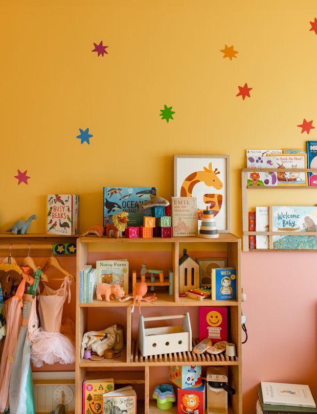 A colourful children's playroom or nursery corner with warm mustard-yellow and pink walls, decorated with playful star decals. Wooden open shelving is filled with children's books, toys, craft items, dolls, and artwork, creating a cheerful, creative, storybook-like atmosphere. A small hanging clothing rail and children's garments are visible at the left.